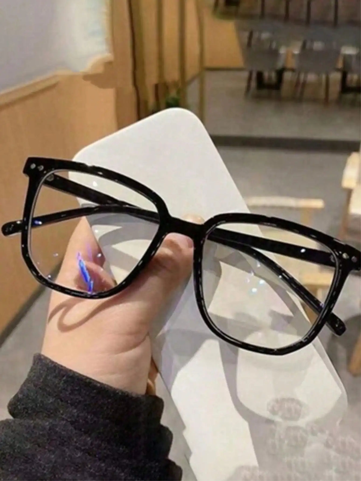 A hand holds a pair of classic black square-shaped eyeglasses against a white surface.