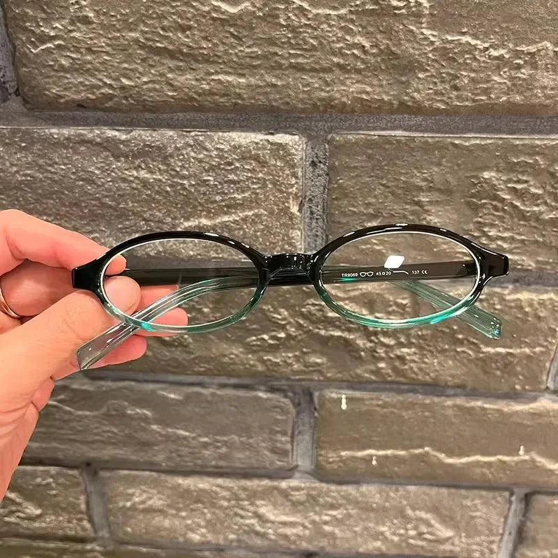 Une main tient une paire de lunettes ovales à monture noire et verres clairs sur un fond de briques grises.