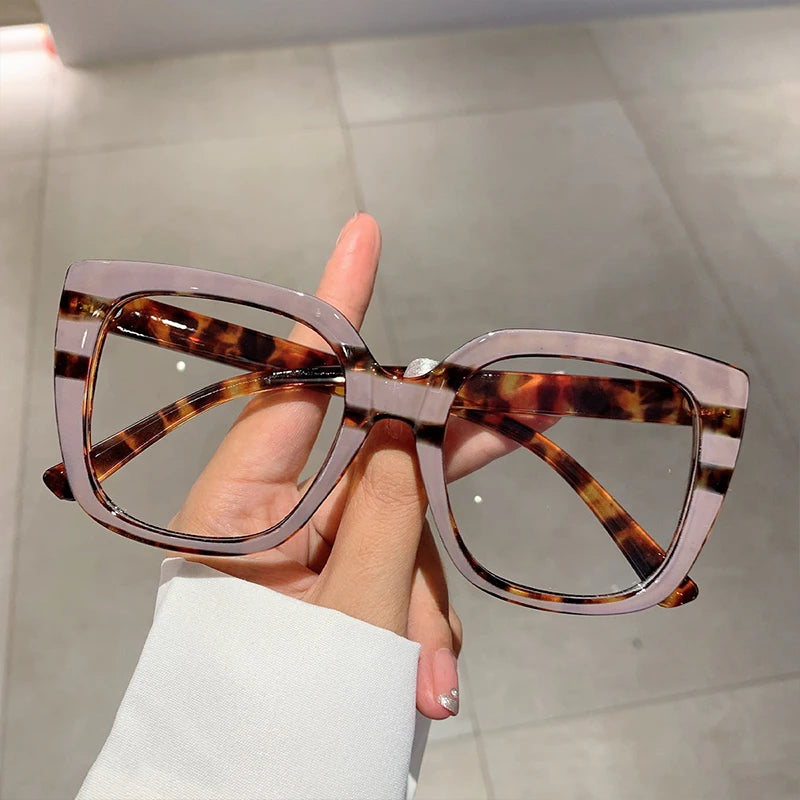 Eine Hand hält eine modische, große quadratische Brille mit braun-schwarz gepunkteten Linsen.