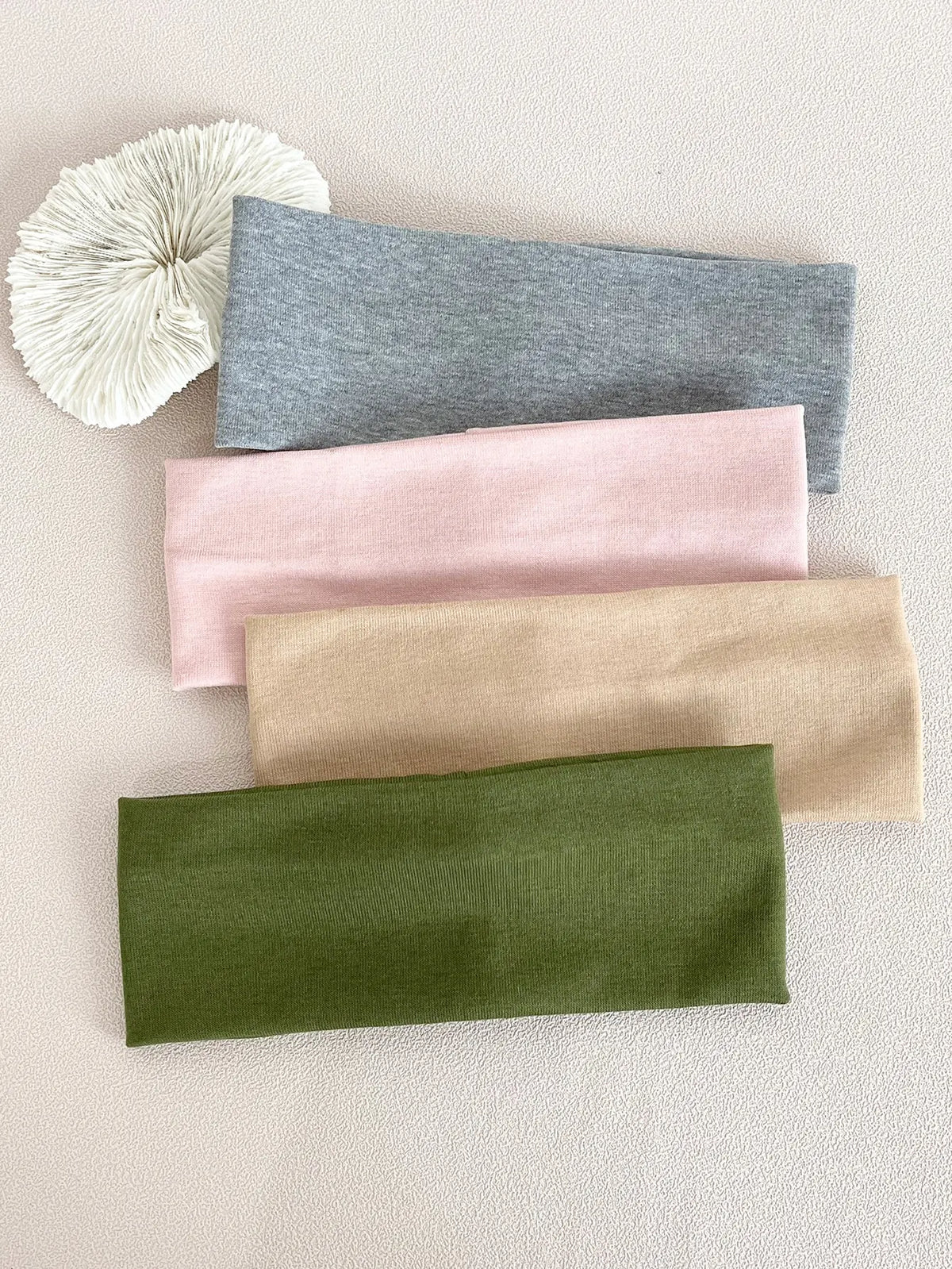 A set of four knitted hair bands in shades of gray, pink, beige, and olive green laid on a light background.