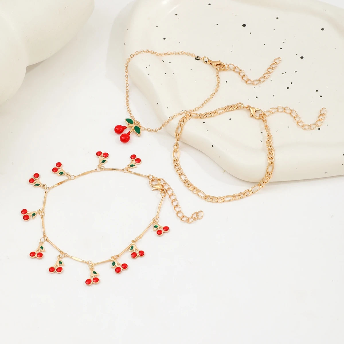 A set of gold-colored anklets featuring small red cherry beads and gold chains displayed on a white surface.