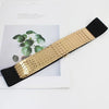 Ceinture élastique large noir et or présentée sur du papier blanc à côté d'une plante verte.