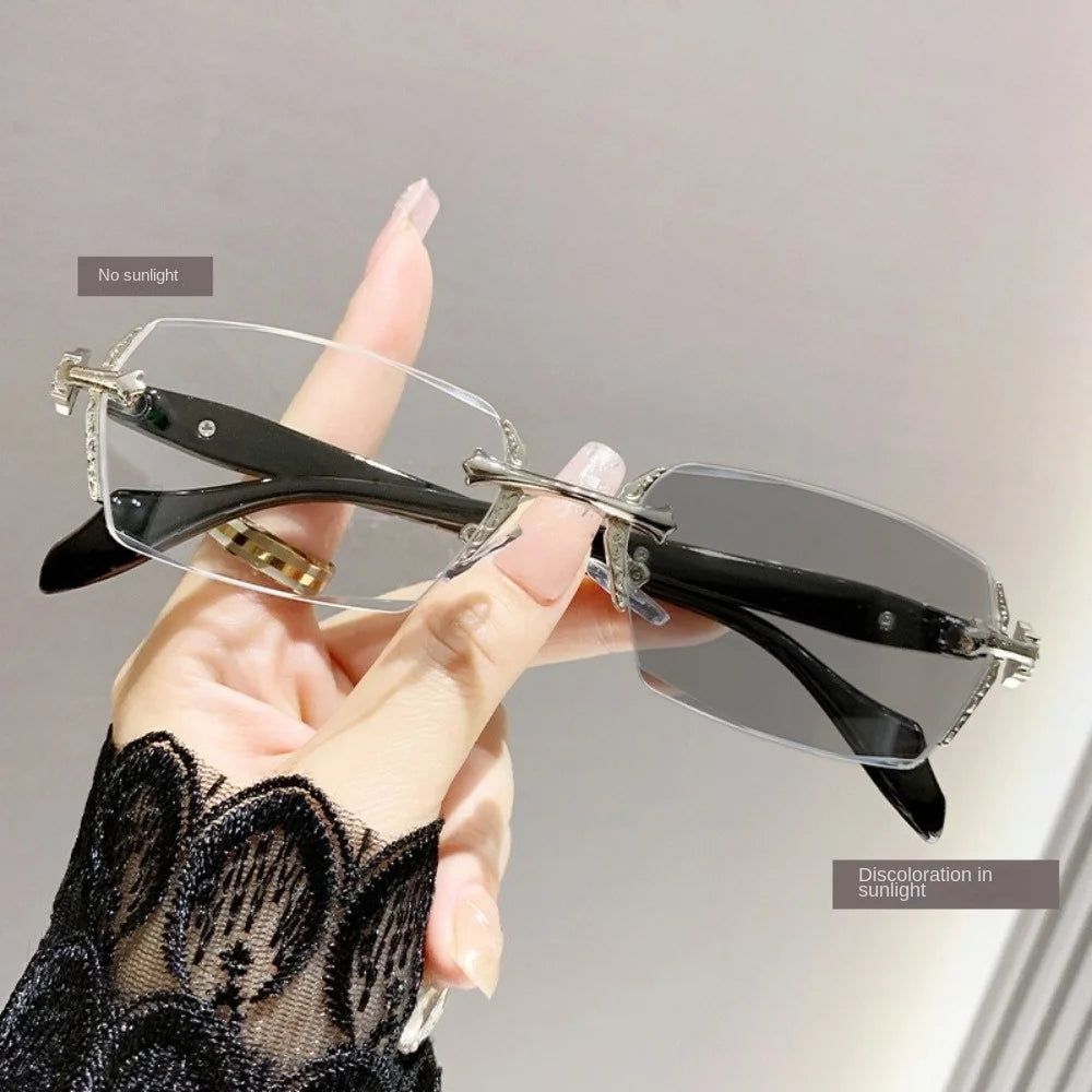 A hand holds a pair of black frameless glasses with clear lenses against a light gray background.