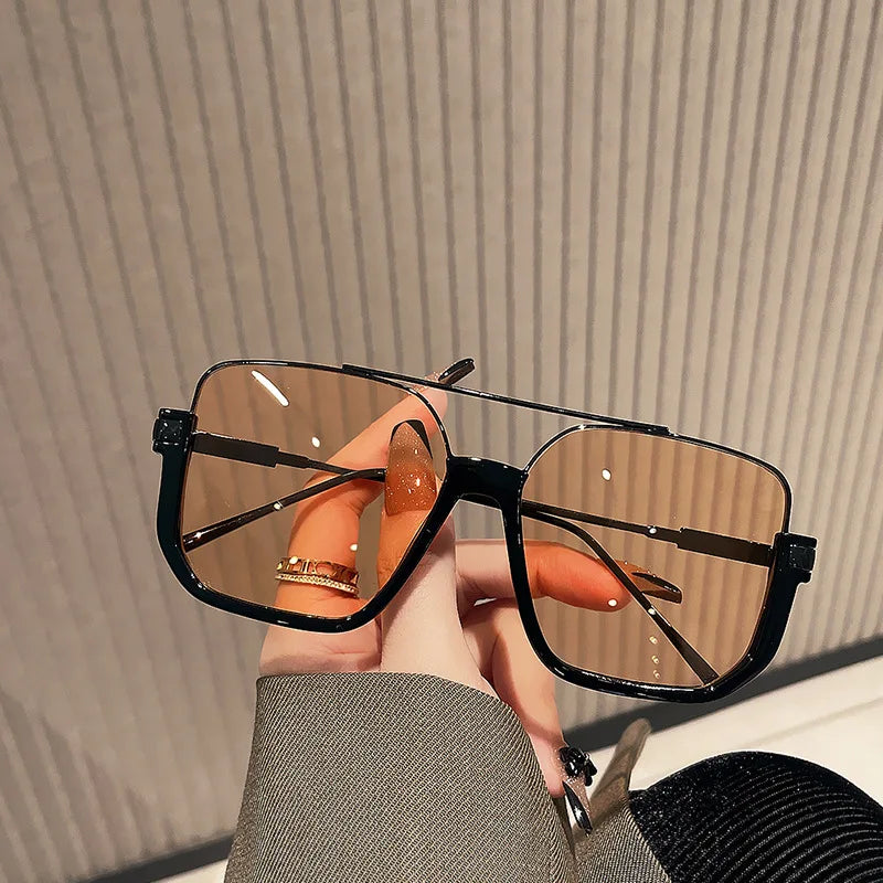 A hand holds a pair of black double bridge oversized square sunglasses with brown lenses against a light background.
