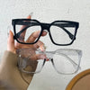 Una mano sostiene un par de gafas cuadradas negras y un par de gafas rectangulares transparentes.