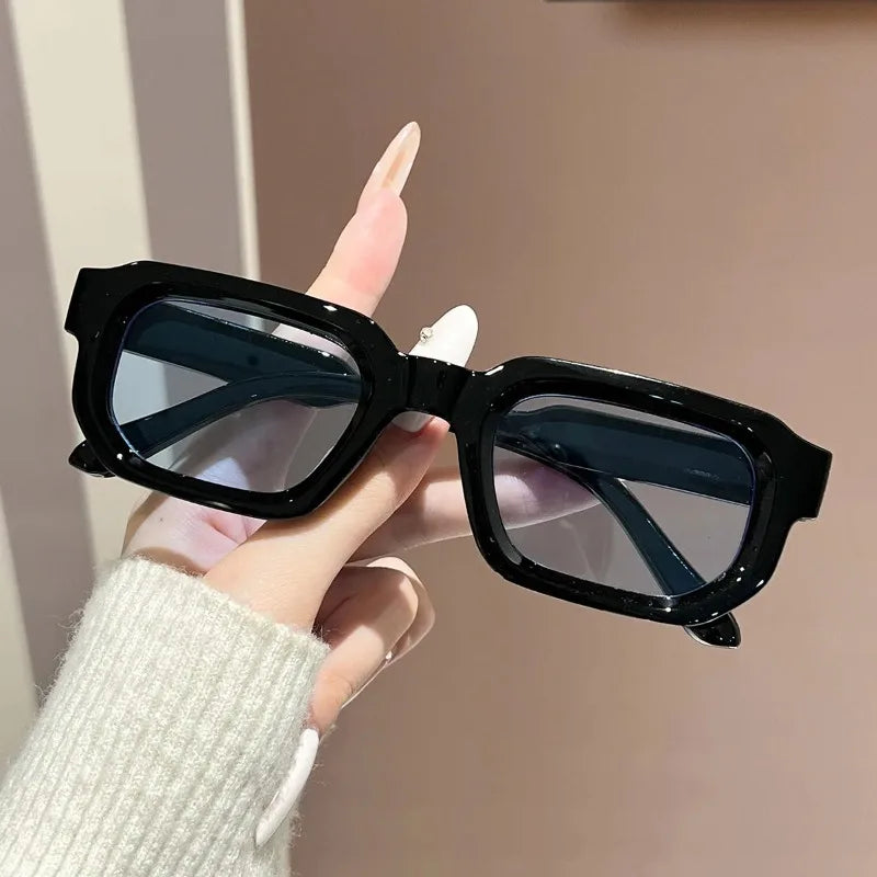 A hand holds a pair of black retro square fashion sunglasses with dark lenses against a light background.