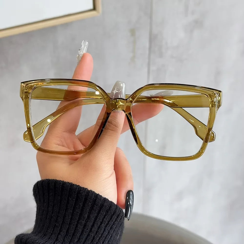 A hand holds a pair of elegant geometric reading glasses with transparent lenses against a light gray background.