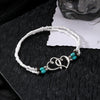Un brazalete de cuentas blanco con pequeños collares de corazón de turquesa y cobre sobre una superficie oscura.