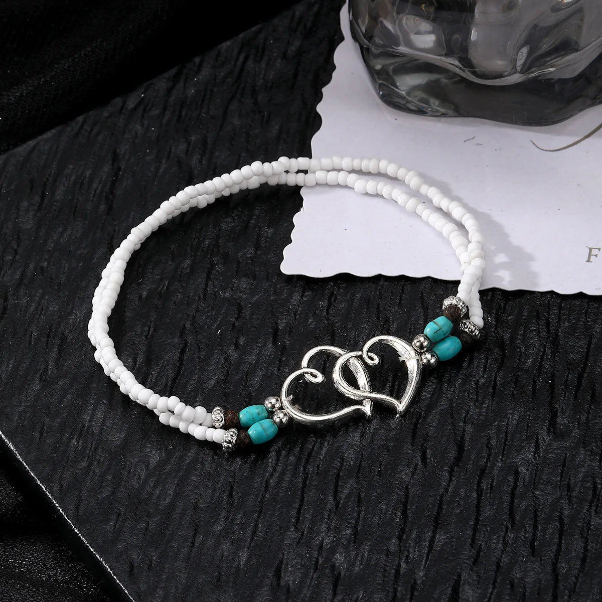 Un brazalete de cuentas blanco con pequeños collares de corazón de turquesa y cobre sobre una superficie oscura.