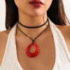 A close-up of a woman wearing a black rope choker necklace with a red hoop pendant against a white top.