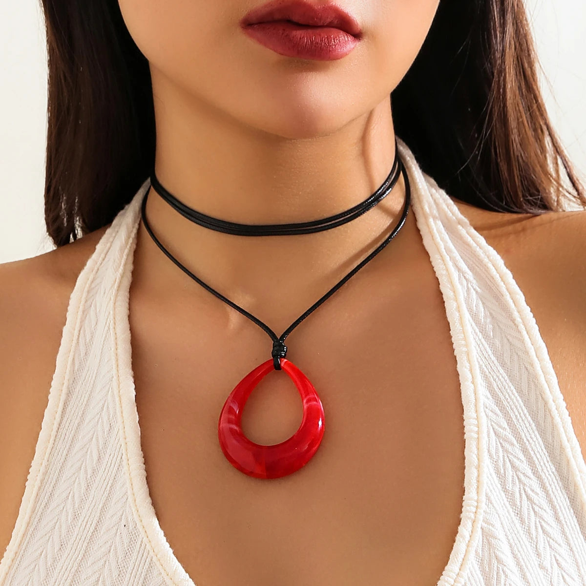 A close-up of a woman wearing a black rope choker necklace with a red hoop pendant against a white top.