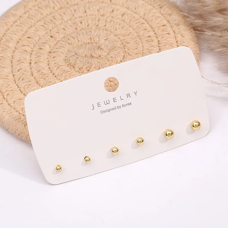 A display of three gold-toned round stud earrings on a white card against a beige fuzzy background.