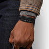 Nahaufnahme eines Mannes Handgelenks mit schwarzem geflochtenem Lederarmband vor dunkelblauer Jeans und einem gemusterten Swe