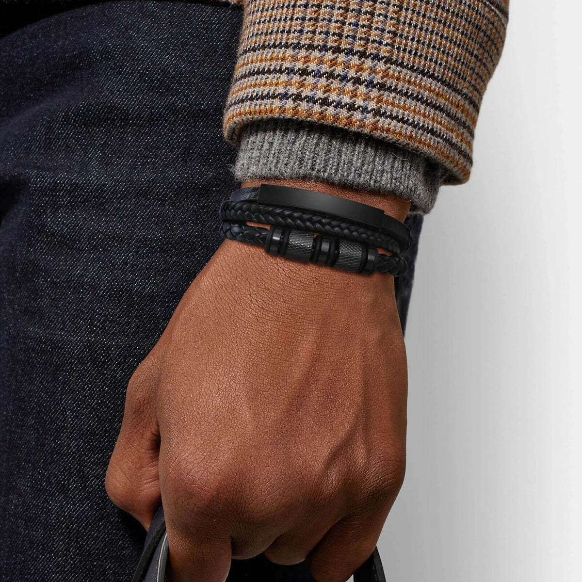 Nahaufnahme eines Mannes Handgelenks mit schwarzem geflochtenem Lederarmband vor dunkelblauer Jeans und einem gemusterten Swe