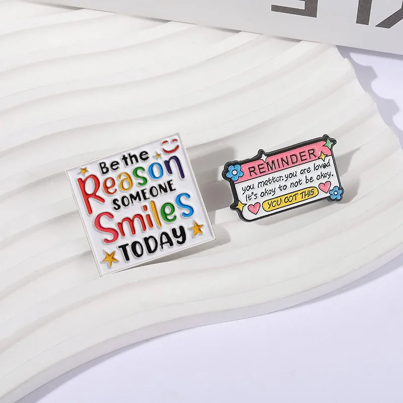 A set of colorful enamel lapel pins featuring text like Be the Reason Someone Smiles Today and a reminder message