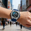 A close-up of a blue sports watch with a metal band worn on a person's wrist in an outdoor city setting.