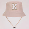 A light beige sun hat with a gold letter K embroidered on the front and a light tan strap.