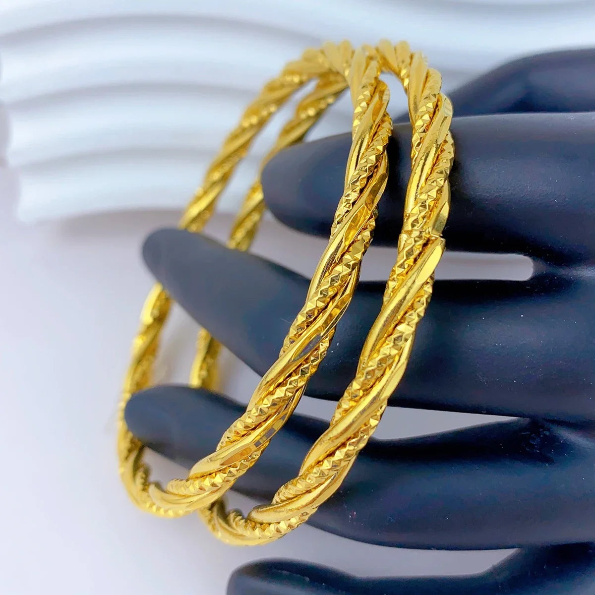 Zwei goldfarbene geometrische Armreifen werden auf einer dunklen Hand präsentiert.