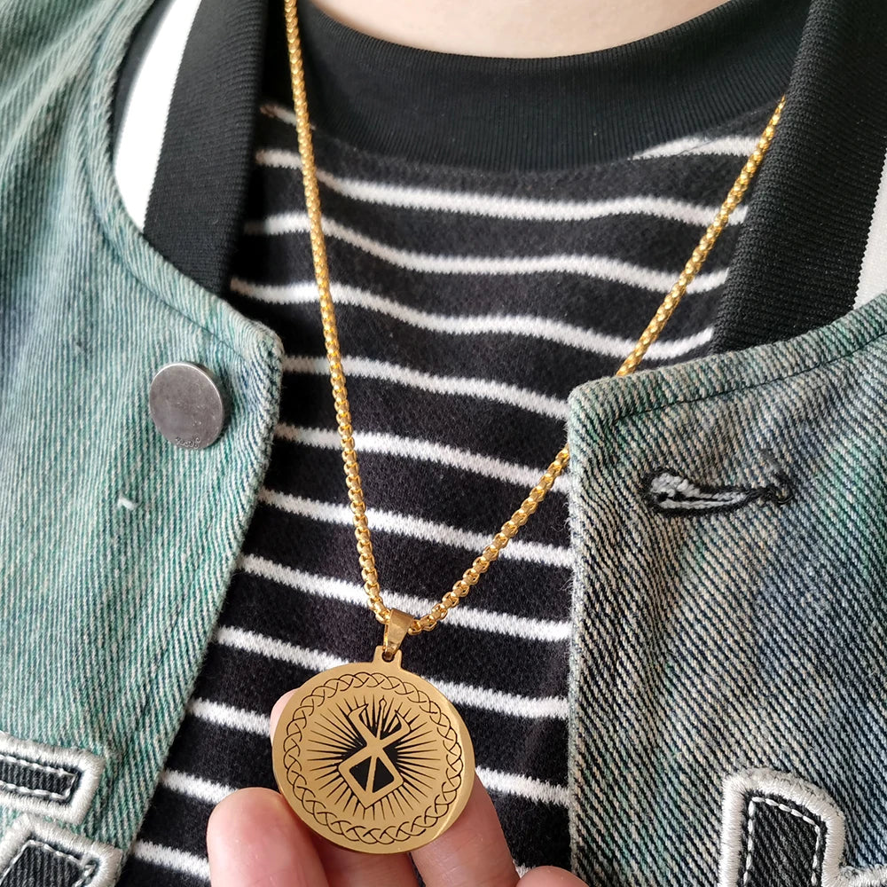 Primer plano de un collar de oro con dije de espada Berserk de anime, usado sobre una camisa a rayas y una chaqueta verde.