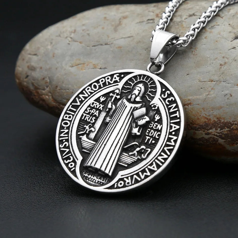 Silver pendant necklace featuring a detailed Saint Benedict exorcism design on a round disc, resting on a smooth stone.