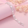 A delicate silver heart charm bracelet is displayed on a pink background next to soft peach-colored leaves.