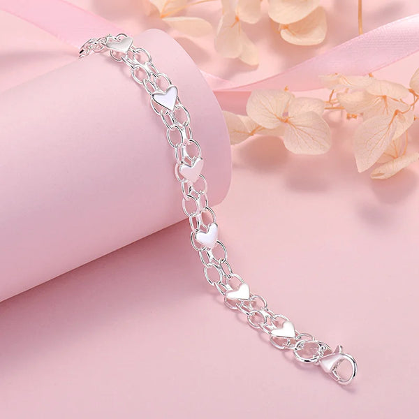 A delicate silver heart charm bracelet is displayed on a pink background next to soft peach-colored leaves.