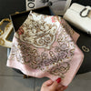 A pink silk scarf with intricate gold and brown floral embroidery is displayed next to a black box and a white clutch.