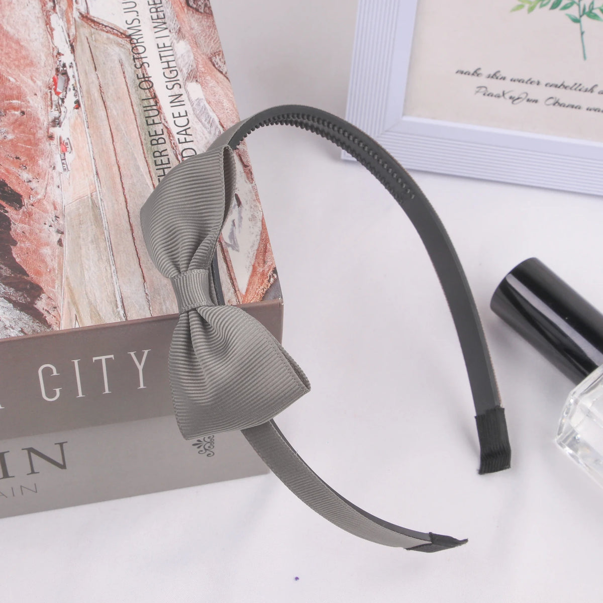 A black grosgrain ribbon hair bow is displayed next to a cosmetic product and a book.