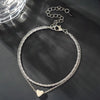 A delicate silver-toned chain anklet with a heart charm is displayed against a dark background.