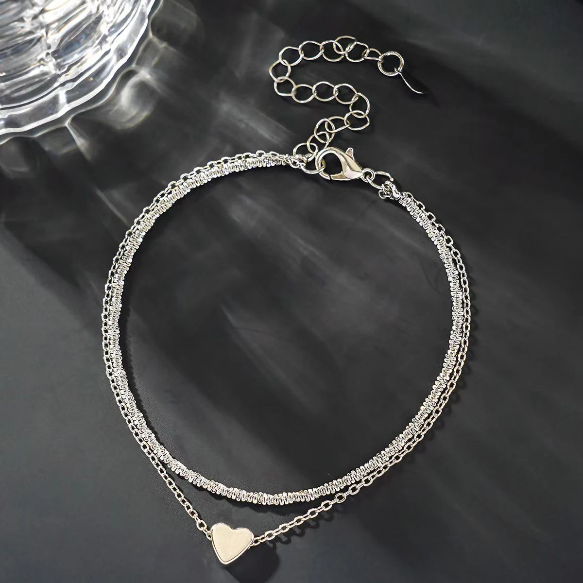 A delicate silver-toned chain anklet with a heart charm is displayed against a dark background.