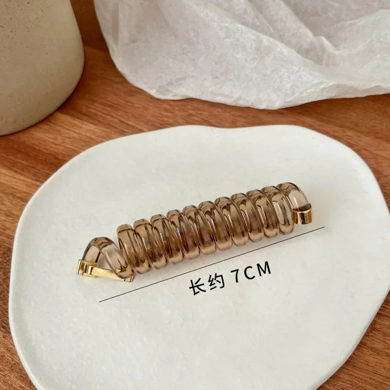 A gold-colored rubber braided ponytail holder is displayed on a white plate, with a measurement line indicating its length of