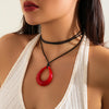 A close-up of a woman wearing a black rope choker necklace with a red hoop pendant against a white top.