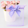 A purple organza drawstring gift bag tied with a lavender ribbon, filled with pink and white flowers.