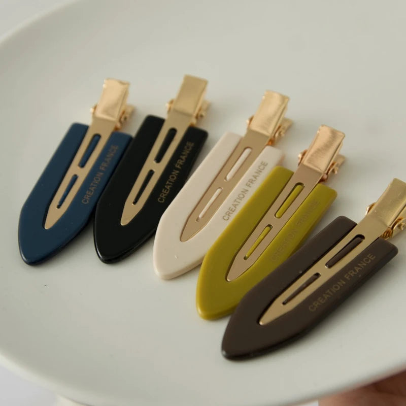 Collection de pinces pour cheveux imprimées en or, dans diverses couleurs (marine, noire, crème, vert olive), présentées sur 