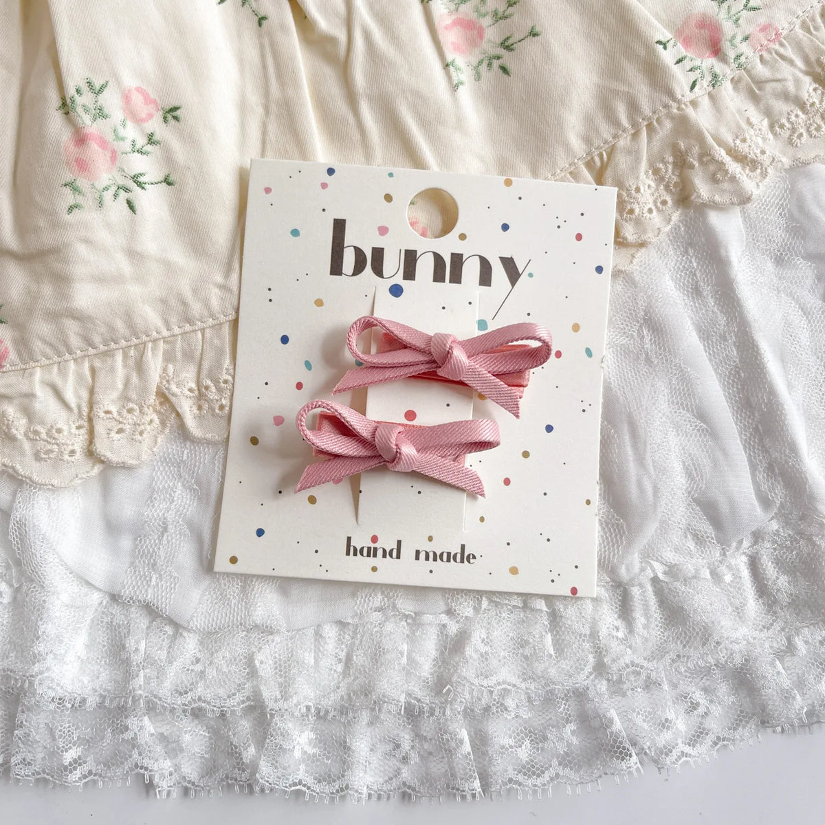 Un set de pinces à cheveux en nœud rose faites à la main, présentées sur une carte avec un fond de tissu en dentelle blanche.