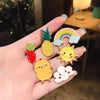 A hand displays several cute animal and plant metal pin badges including a pineapple, a strawberry, a rainbow, a sun, and a c