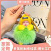 A hand holds a bright green fluffy plush doll with yellow accents and a knitted hood, resembling a cartoon character keychain