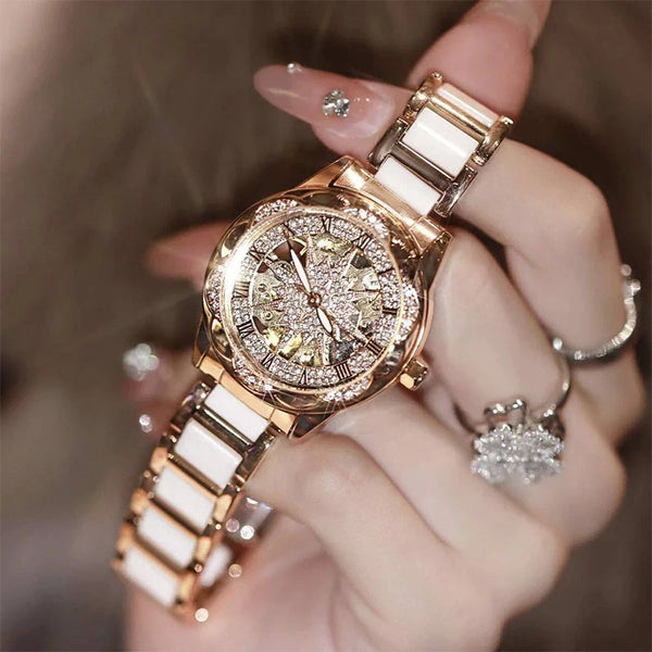 A hand is holding a gold-toned quartz diamond wristwatch with a white strap against a blurred background.