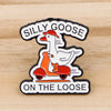 An enamel pin featuring a cartoon goose riding a scooter with the text Silly Goose On The Loose