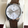 Montre argentée avec cadran blanc et chiffres romains affichée sur une boîte blanche.