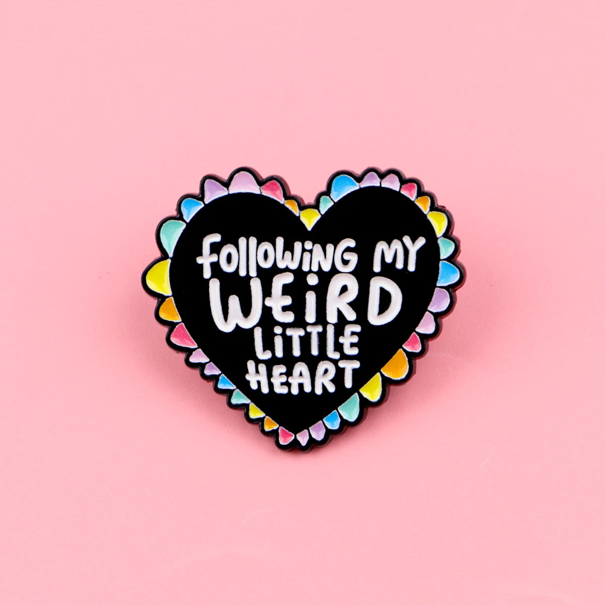 A black heart enamel pin with the text Following my weird little heart surrounded by colorful ruffled edges.
