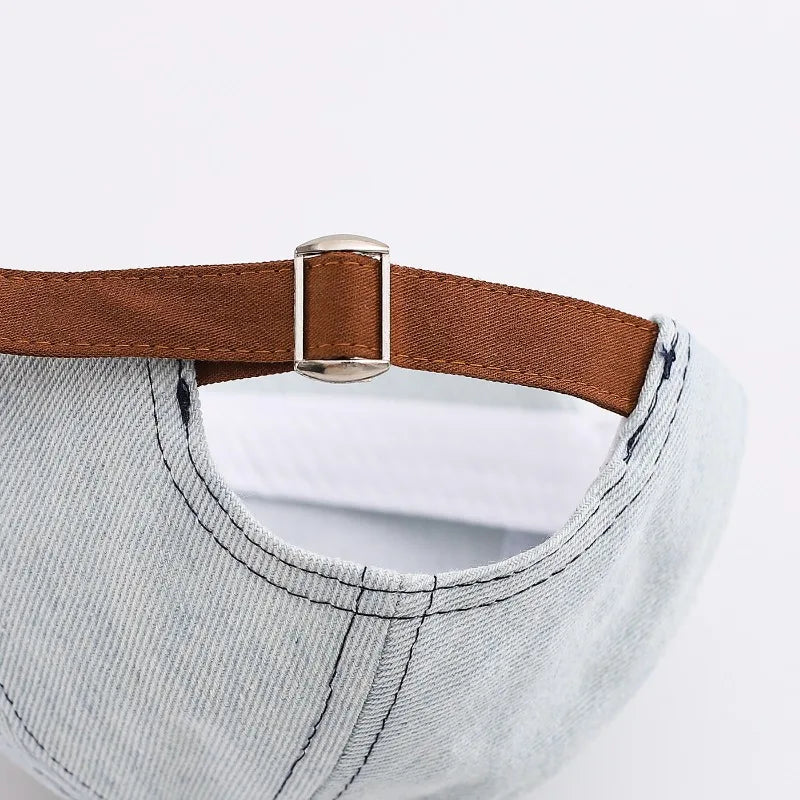 A close-up of a light gray denim baseball cap with a brown leather strap and silver buckle.