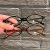 Une main tient une paire de lunettes à ovales rétro brun-tintées sur un fond de briques grises.