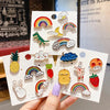 A hand holds several colorful metal pin badges featuring rainbows, clouds, and cute animals like dinosaurs, pandas, and rabbi
