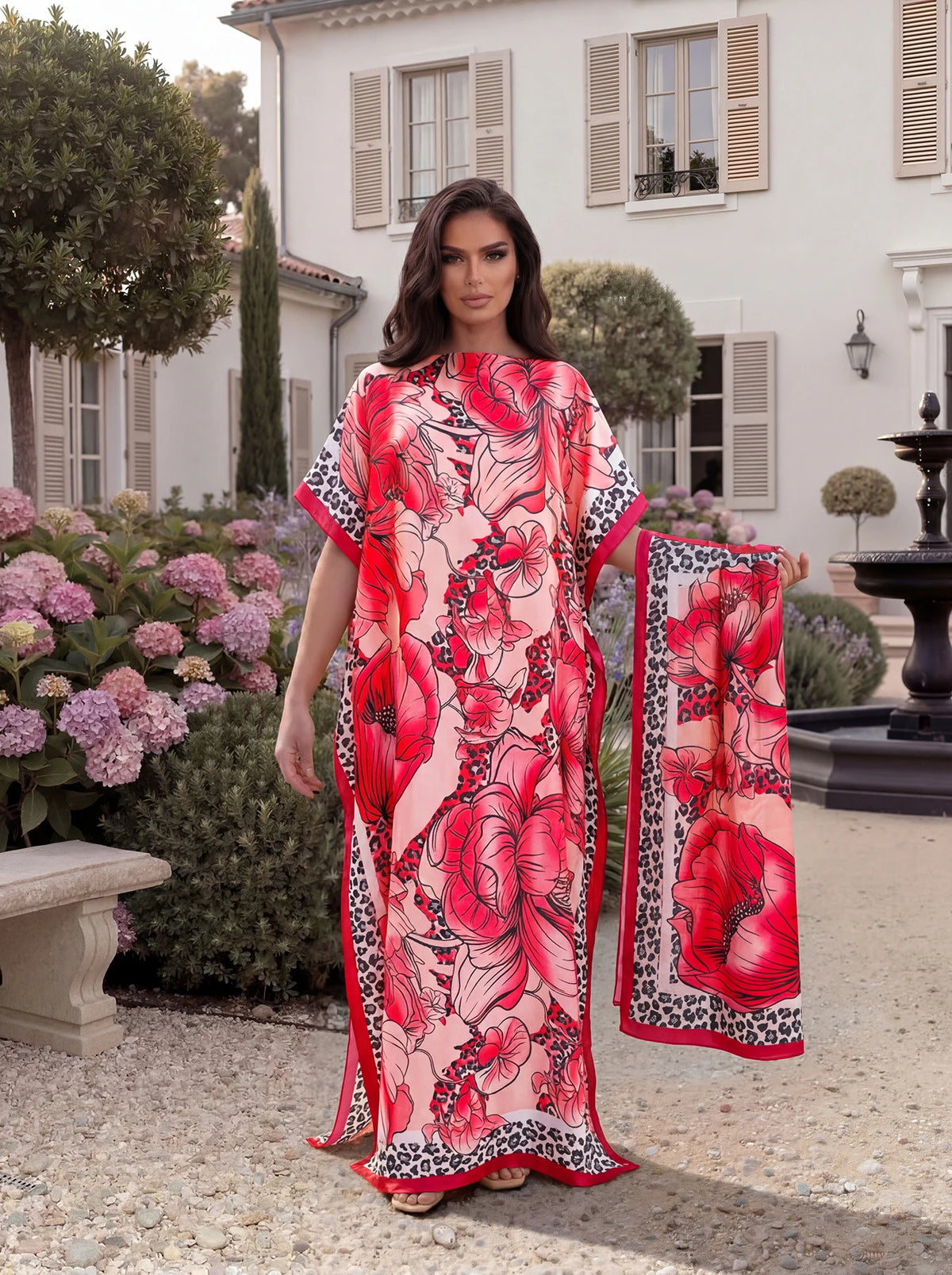 A woman models a pink and red tropical floral leopard print abaya scarf outdoors in a garden setting.
