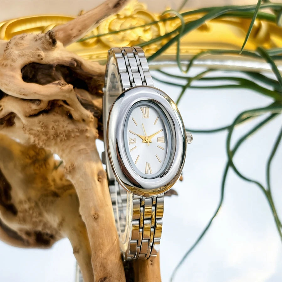 Reloj vintage con correa de acero plateado y esfera blanca, exhibido contra un fondo de ramas secas y follaje verde