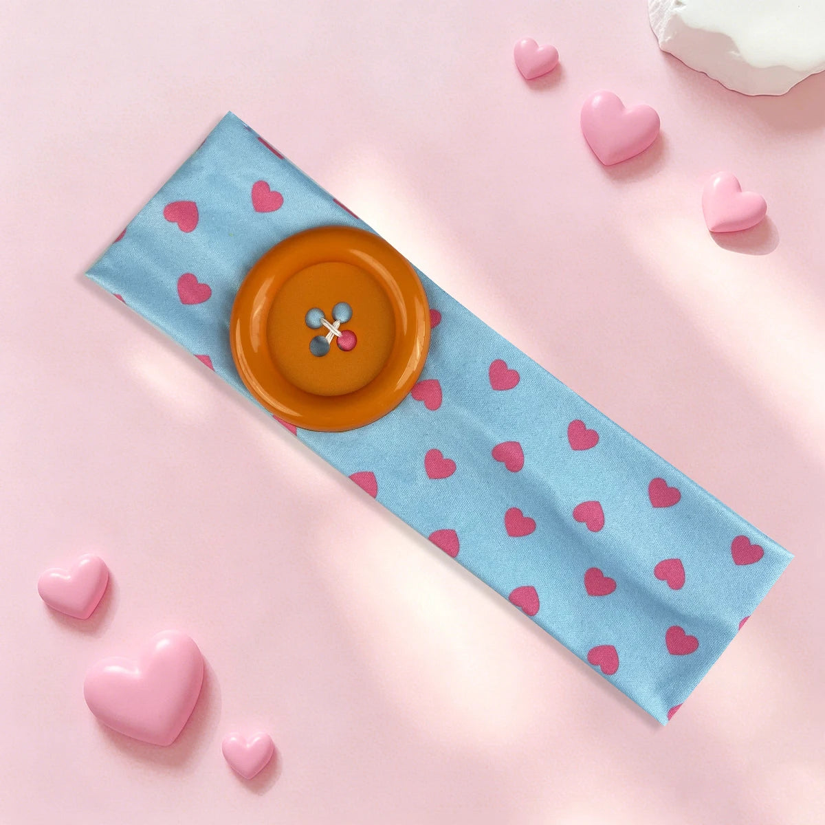 A light blue headband with pink hearts and an orange button, lying on a pink surface.