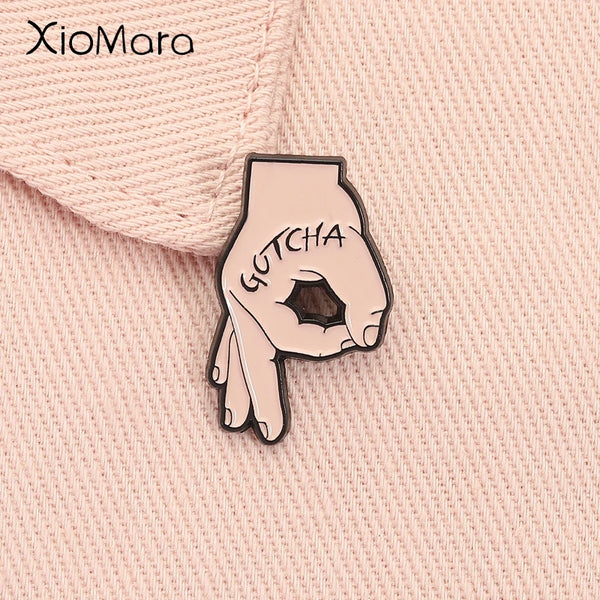 A close-up of a pink knitted fabric with a gold-toned enamel hand gesture brooch featuring the word GOTCHA.