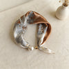 A silk scarf featuring a beige and bronze leaf print tied with a pearl and magnetic buckle.