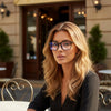 Une femme portant des lunettes transparentes bleues minimalistes de blocage de lumière devant une entrée de bâtiment.